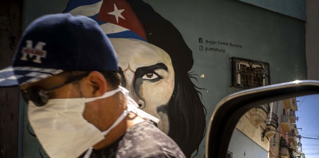 A man wearing a mask walks alongside a mural of Ernesto 